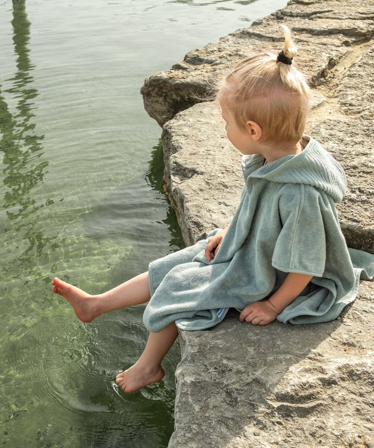 Kind mit Badeponcho sitzt draußen am Badesee.