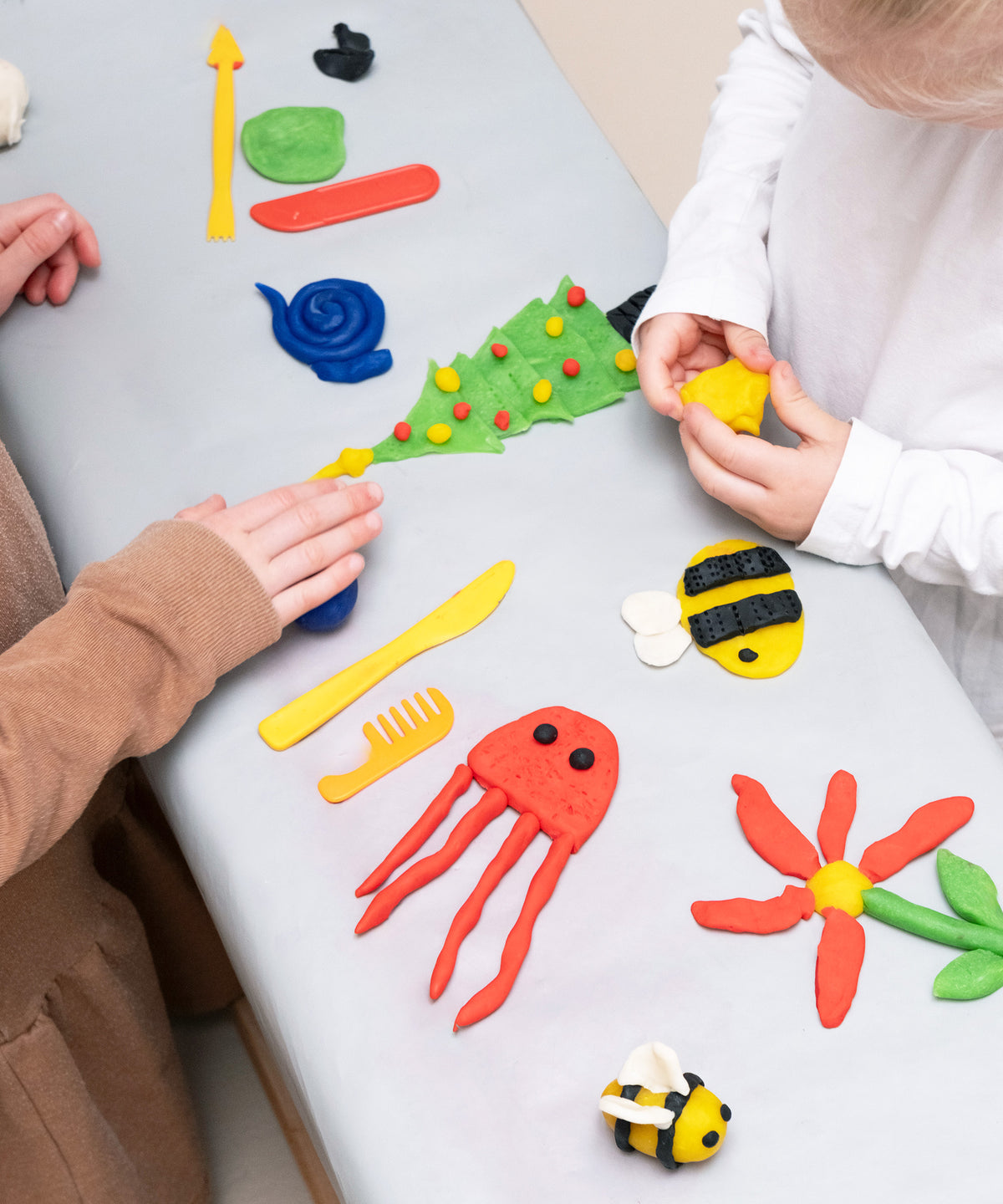 Zwei Kinder spielen mit Knete, verschiedene Figuren liegen auf dem Tisch, z. B. ein Tannenbaum und eine Qualle.