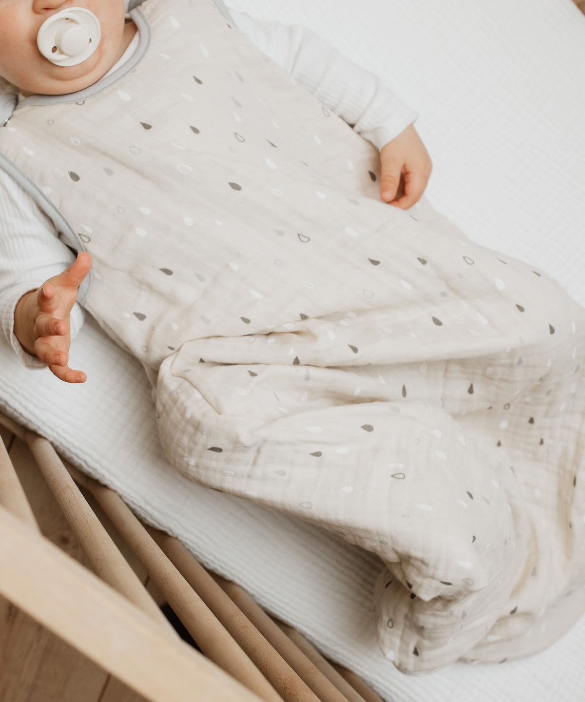 Baby mit Sommerschlafsack Musselin in Natur Tropfen liegt in Bettchen.
