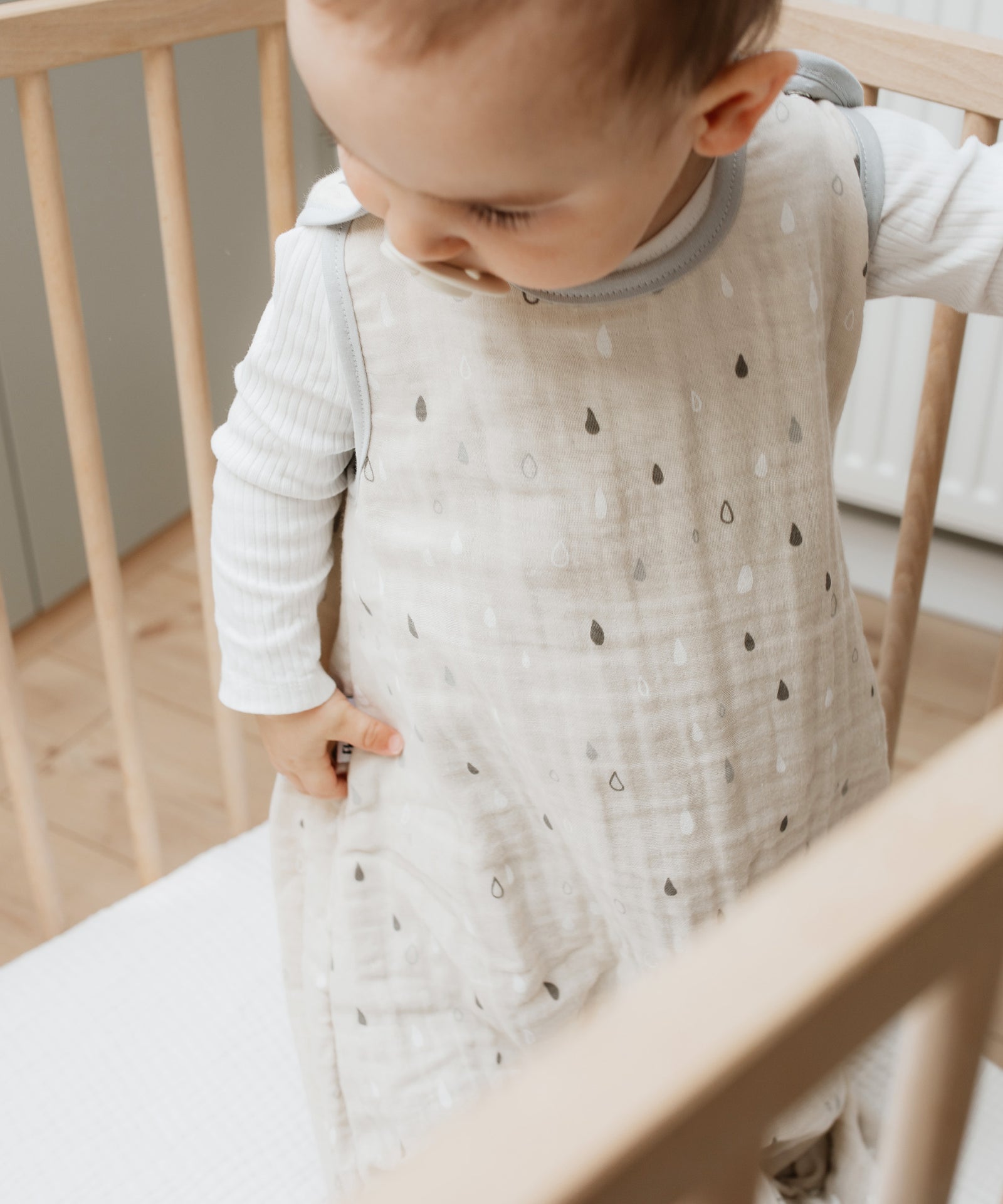 Baby mit Sommerschlafsack aus Musselin steht in Bettchen.