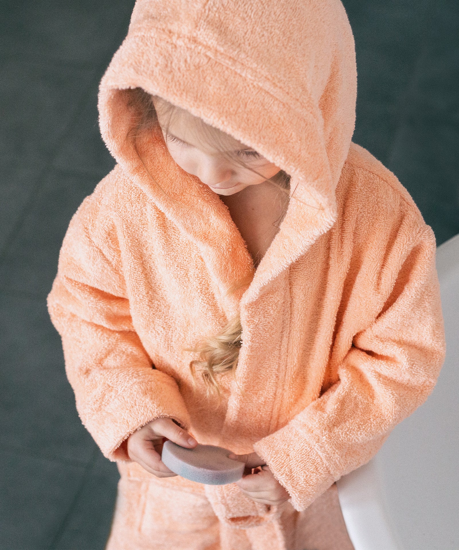 Kind trägt den Bademantel mit aufgesetzter Kapuze und hält einen Schwamm.