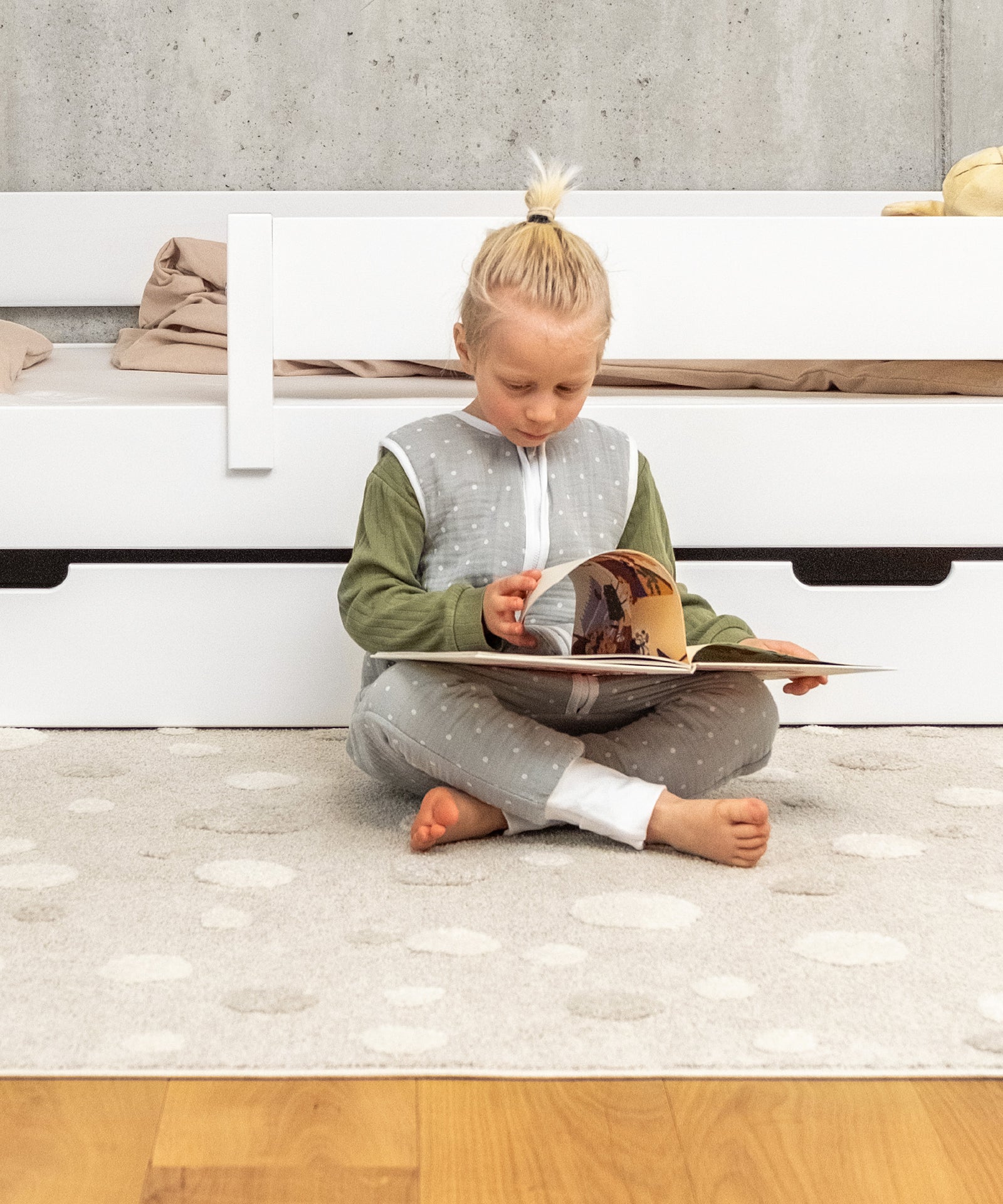 Ein Kind sitzt auf einem Teppich vor einem weißen Hausbett und liest ein Buch.