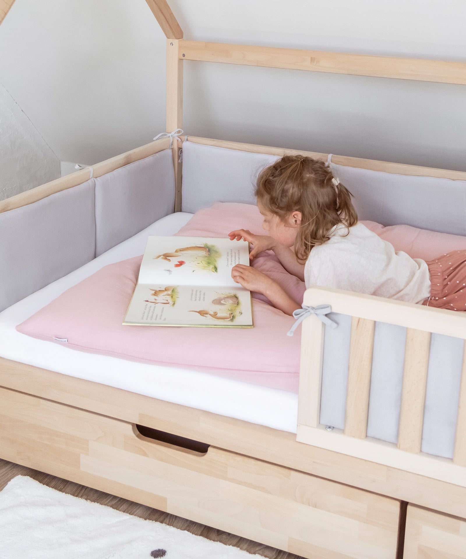 Gemütliche Kuschelecke*Die weich gepolsterte Bettumrandung sorgt für Gemütlichkeit im Bett.|Mädchen liegt in Bett mit Bettumrandung und schaut ein Bilderbuch an.