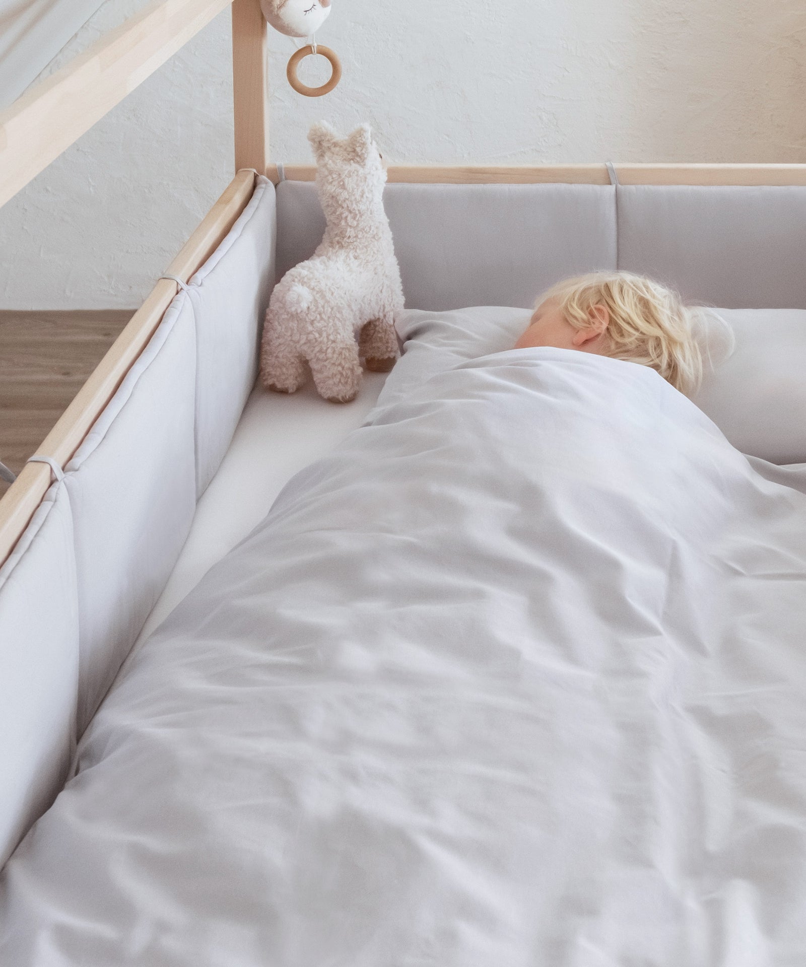 Sicher & geborgen*Extra weicher Schutz für dein Kind durch die weich gefütterte Polsterung rund um das Bett.|Kind liegt in Bett mit Bettumrandung und Bettwäsche aus gleichem Material in Hellgrau und schläft.