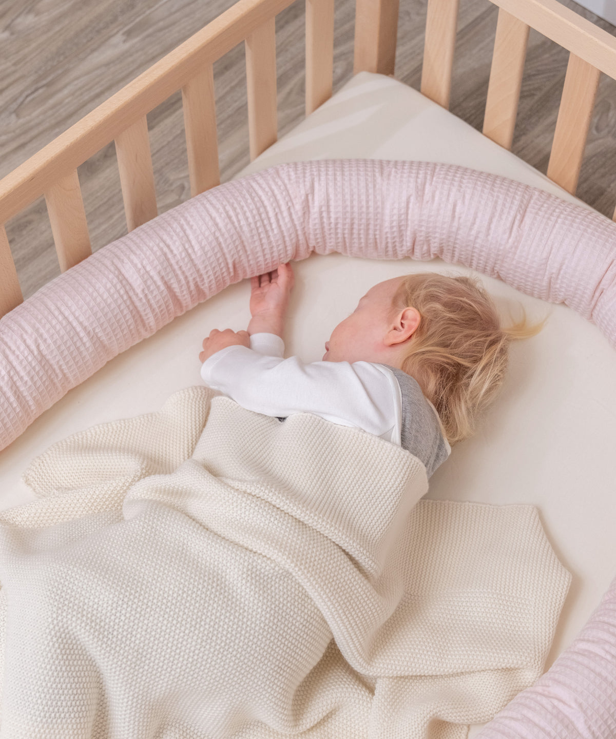 Baby liegt in Bett und schläft, rosa Bettschlange liegt am Gitter entlang.