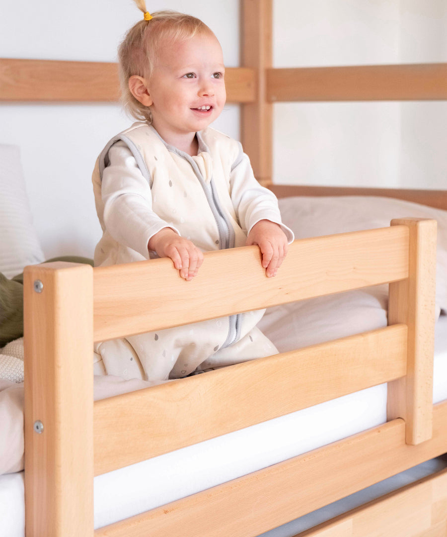 Optionaler Rausfallschutz*Für noch mehr Sicherheit kann unser Rausfallschutz bei Bedarf ganz leicht angebracht werden.|Kind sitzt im Bett und greift an den optionalen Rausfallschutz aus Buchenholz.