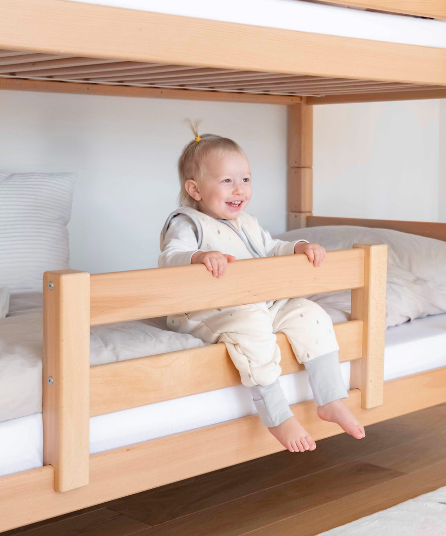 Optionaler Rausfallschutz*Für noch mehr Sicherheit kann unser Rausfallschutz bei Bedarf ganz leicht angebracht werden.|Kind sitzt im Bett und greift an den optionalen Rausfallschutz aus Buchenholz.