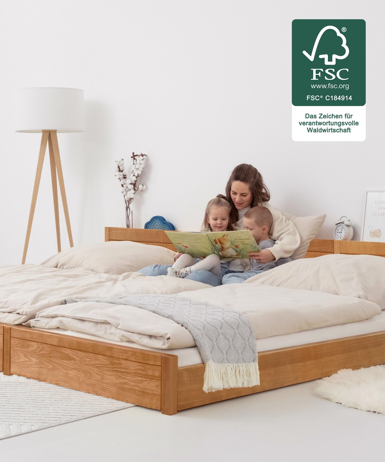 Das perfekte Familienbett*Flexibel dank modularer Bauweise und passend für jedes Zuhause: Unser Bett ist in zwei Höhen und drei Ausführungen erhältlich.|Mutter mit Kindern schaut Bilderbuch an auf Familienbett Esche bodentief.