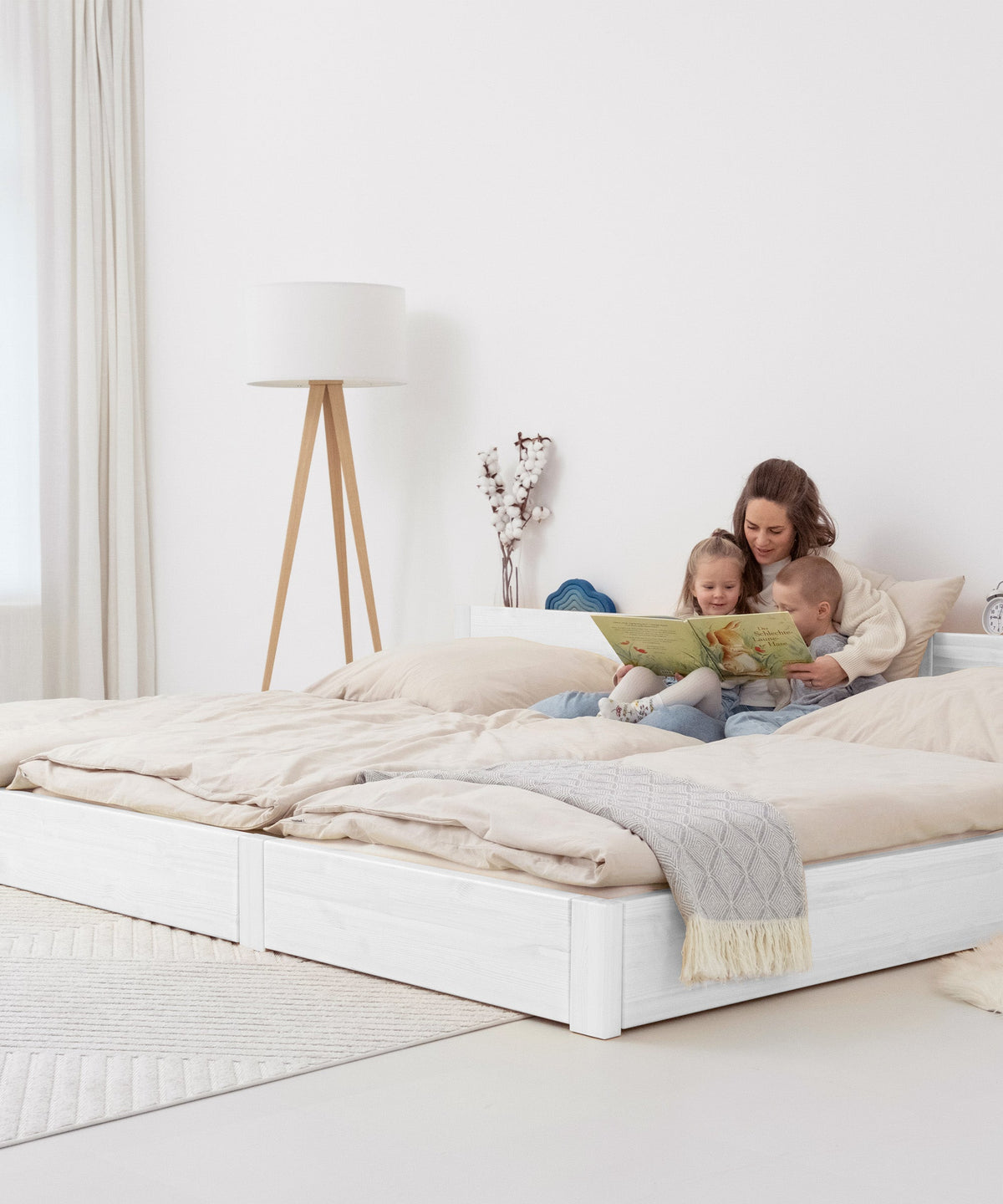 Familie sitzt auf Familienbett weiß bodentief und schaut ein Buch an.