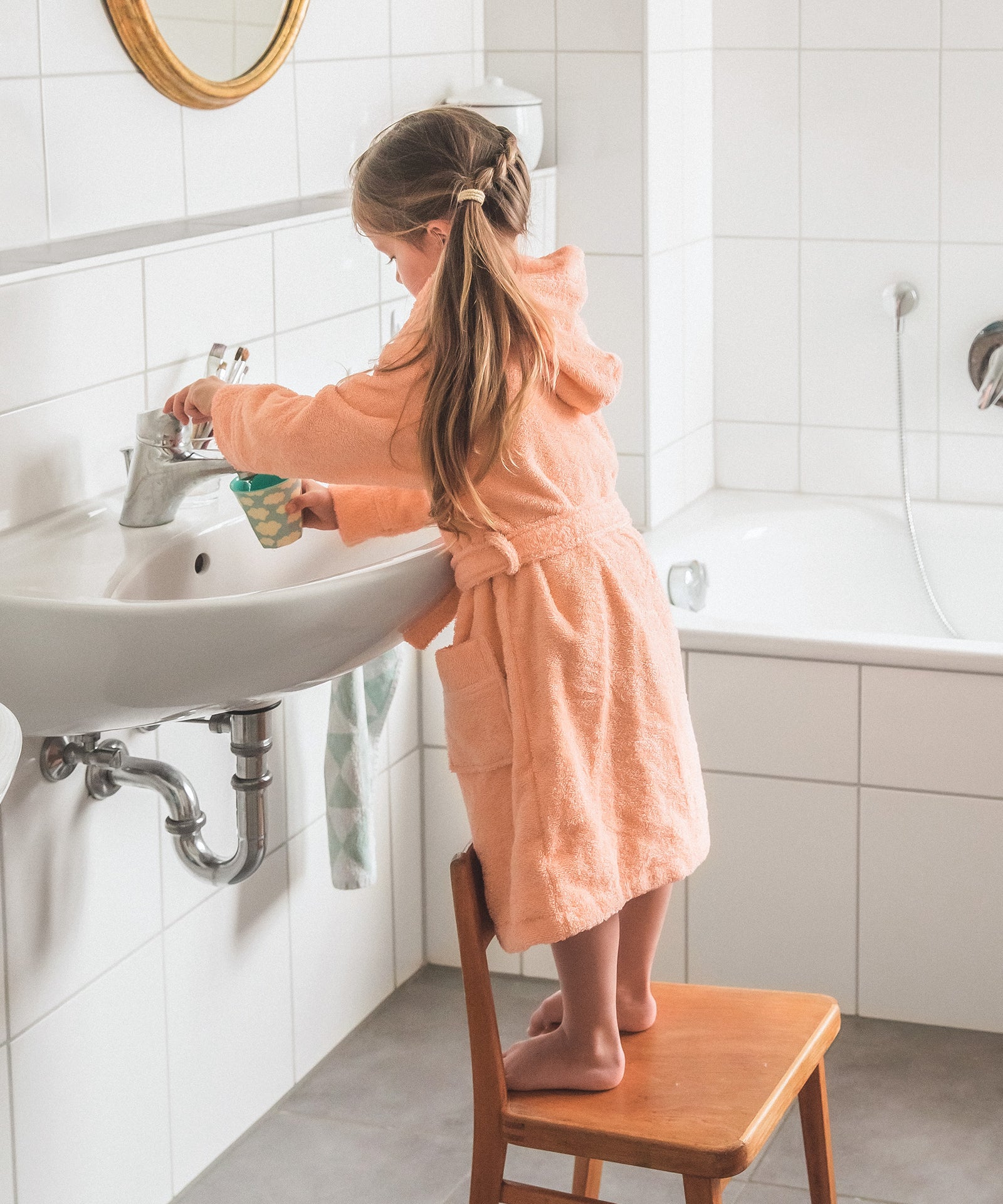 Kind steht auf einem Stuhl vor dem Waschbecken und füllt sich Wasser in einen Becher.