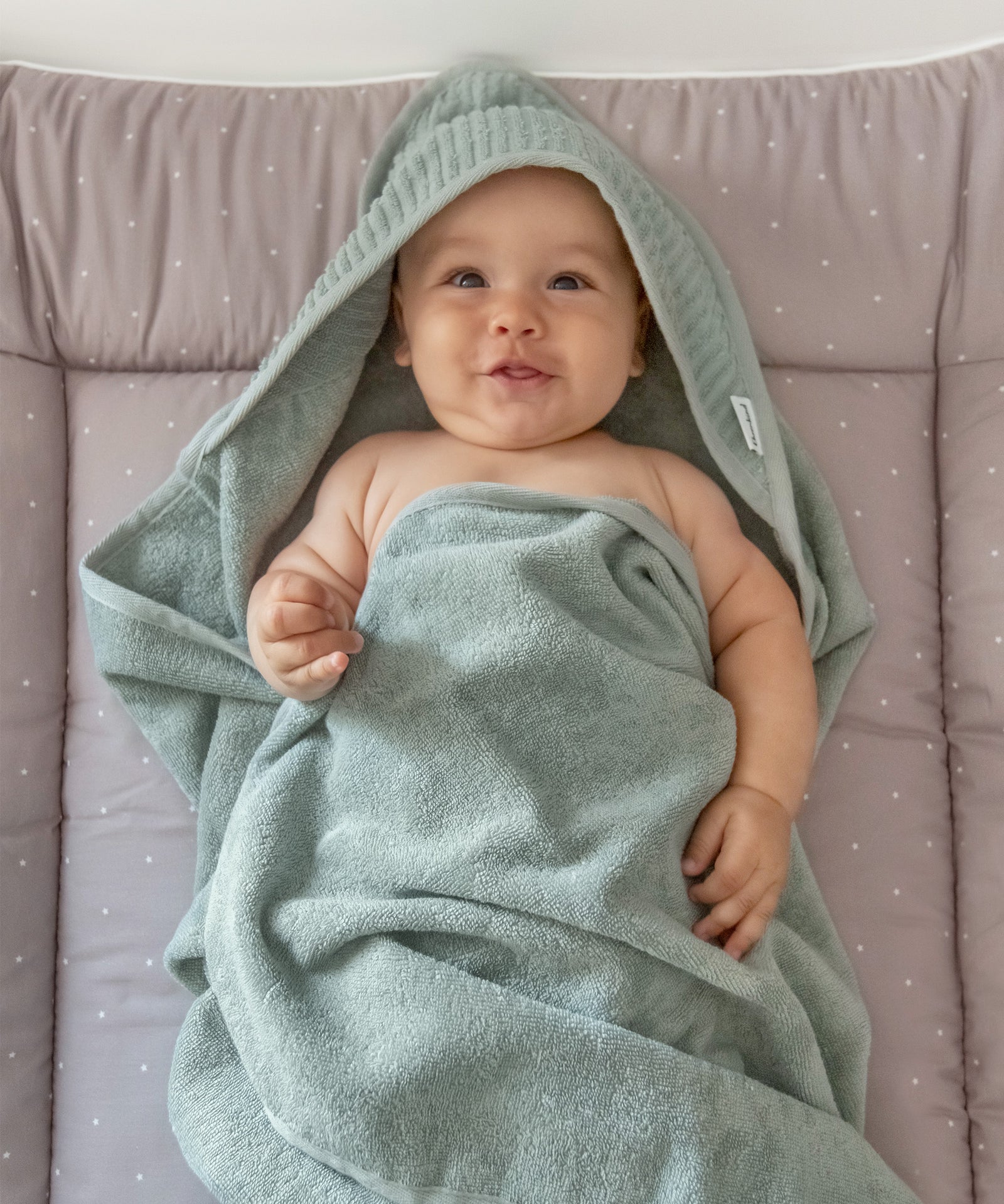 100% Bio-Baumwolle*Ein gutes Gefühl bei empfindlicher Haut - das Kapuzenhandtuch aus reiner Bio-Baumwolle ist OEKO-TEX® STANDARD 100 zertifiziert.|Baby mit Kapuzenhandtuch Jade liegt auf Wickeltisch.