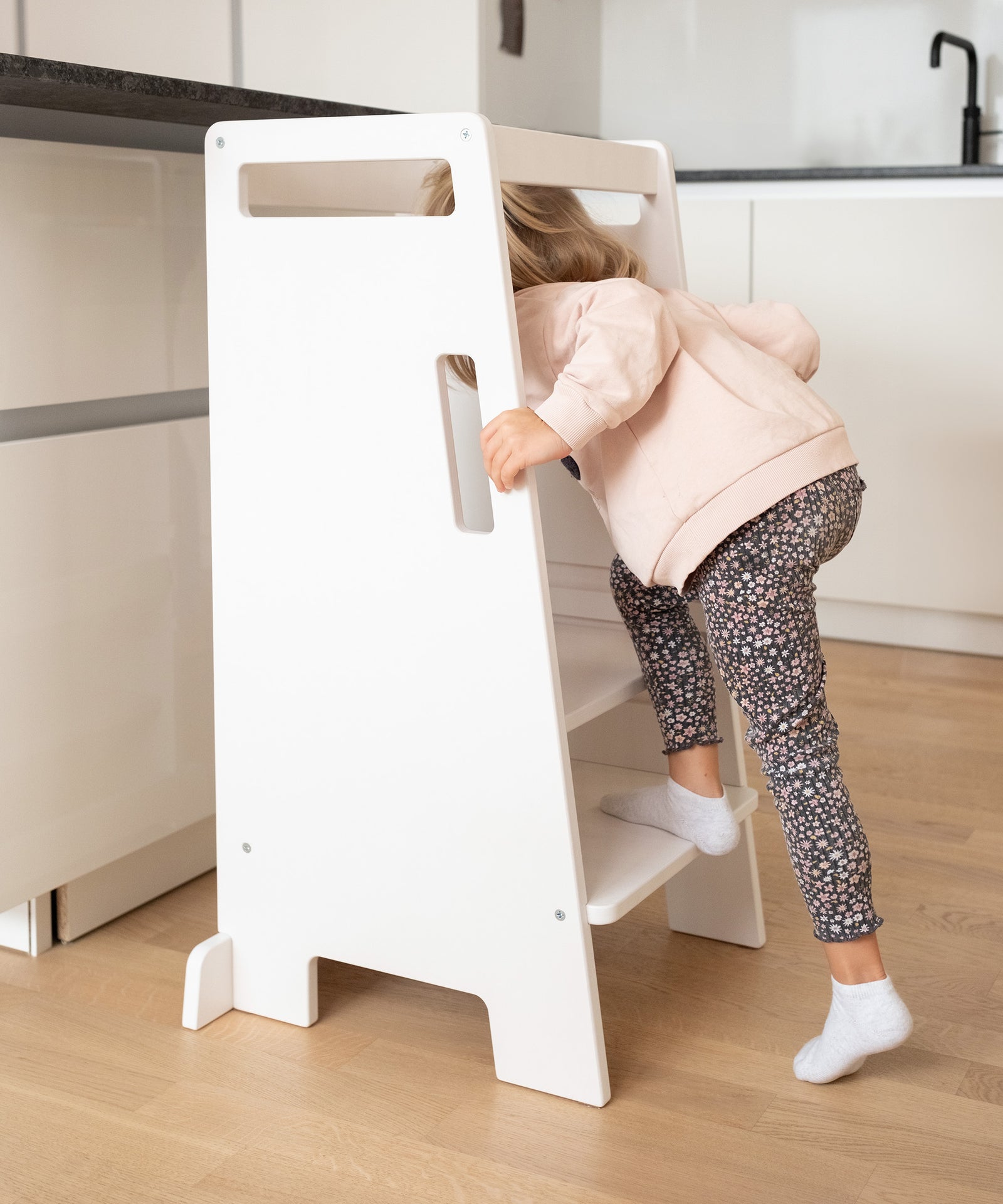 Stabil & kippsicher*Das stabile Design macht den Stufenhocker kippsicher, während Rausfallschutz und Grifflöcher dem Kind sicheren Halt geben.|Ein Kind klettert auf den weißen Lernturm in einer Küche.