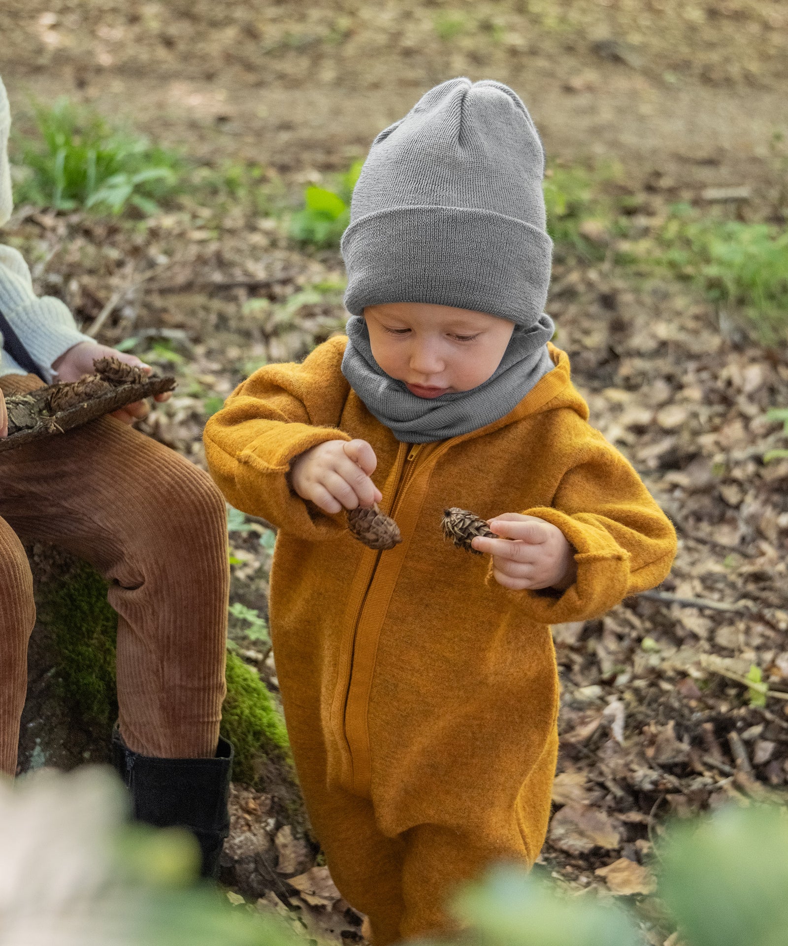 Kleinkind im Walkoverall mit Strickmütze und Schlauchschal in Grau spielt mit Tannenzapfen.
