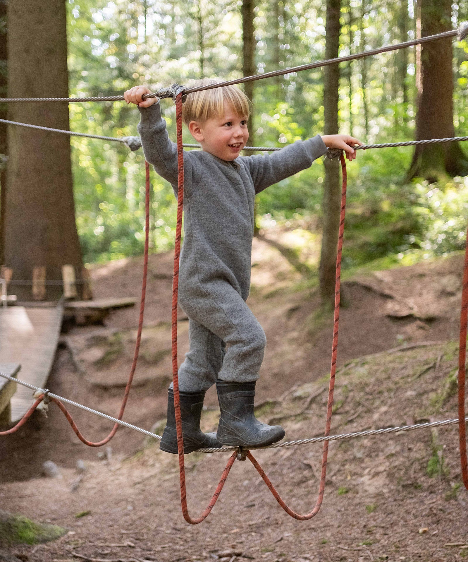 Junge mit Wollfleeceoverall Grau auf Klettergerüst im Wald.