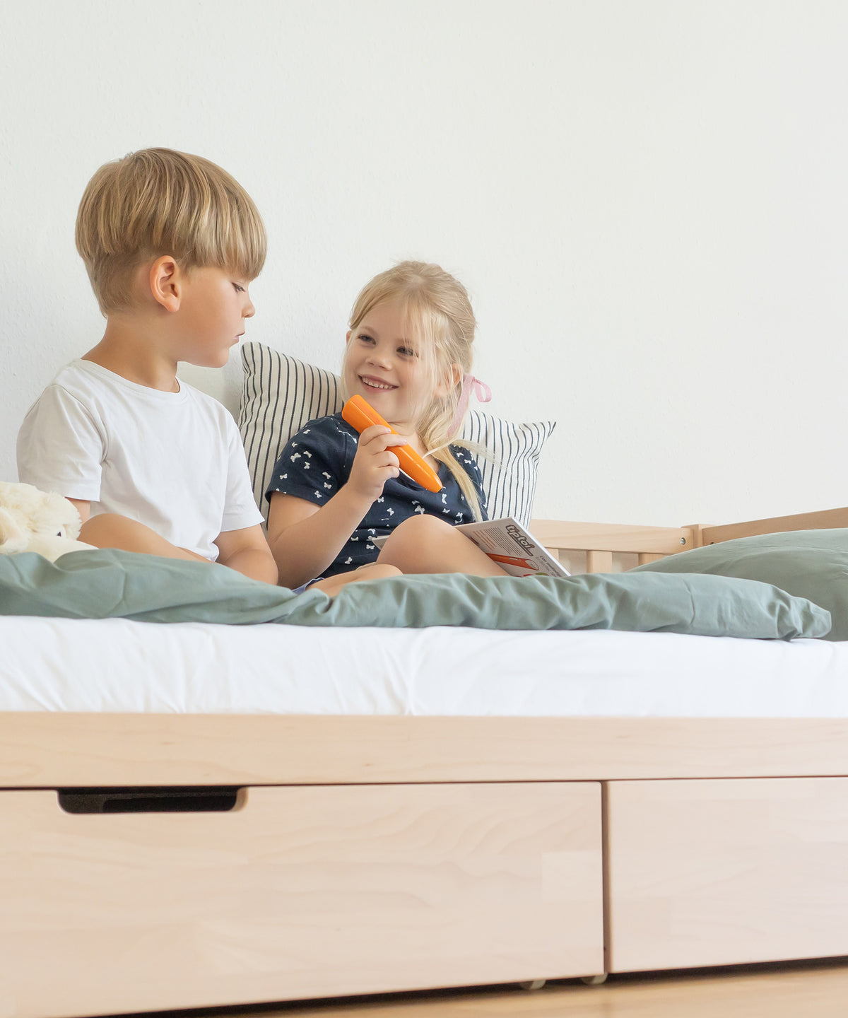 Zwei Kinder sitzen auf Kinderbett Buche natur mit Bettschubladen und schauen Buch an.