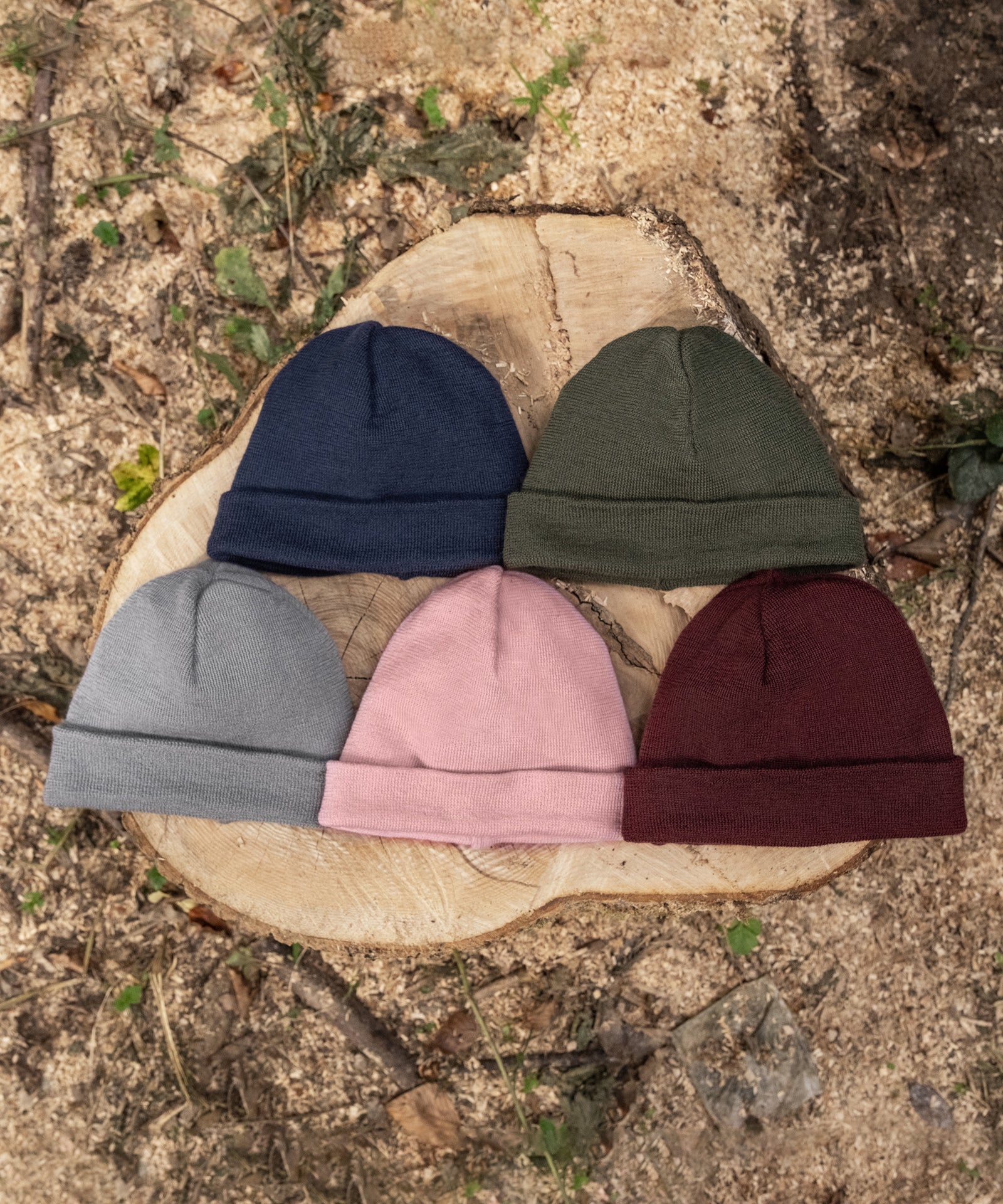 Nachhaltig*Die extrafeine Schurwolle für die Mütze ist mulesingfrei und stammt aus kontrolliert biologischer Tierhaltung (k.b.T.).|Strickmützen in fünf Farben liegen nebeneinander auf einem Baumstumpf im Wald.