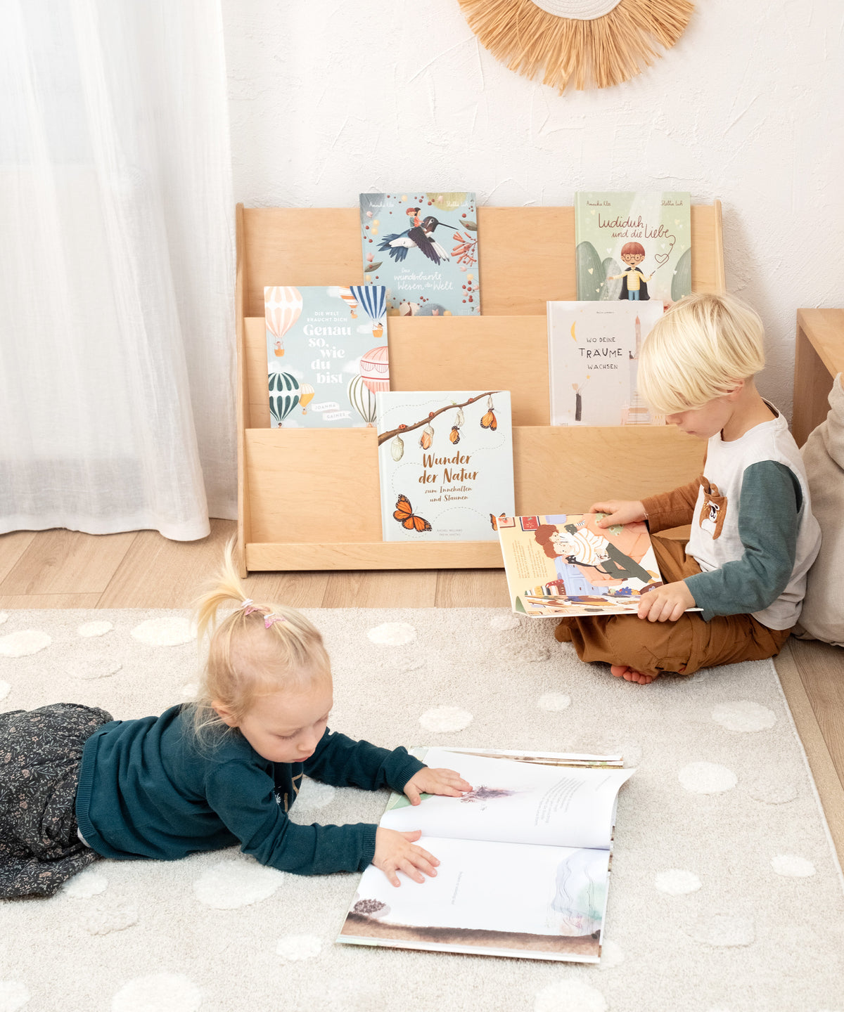 Zwei Kinder sitzen vor dem Bücherregal, das mit Kinderbüchern bestückt ist. Eines der Kinder liest ein Buch auf dem Boden liegend, das andere sitzt auf einem Teppich und blättert ebenfalls in einem Buch.