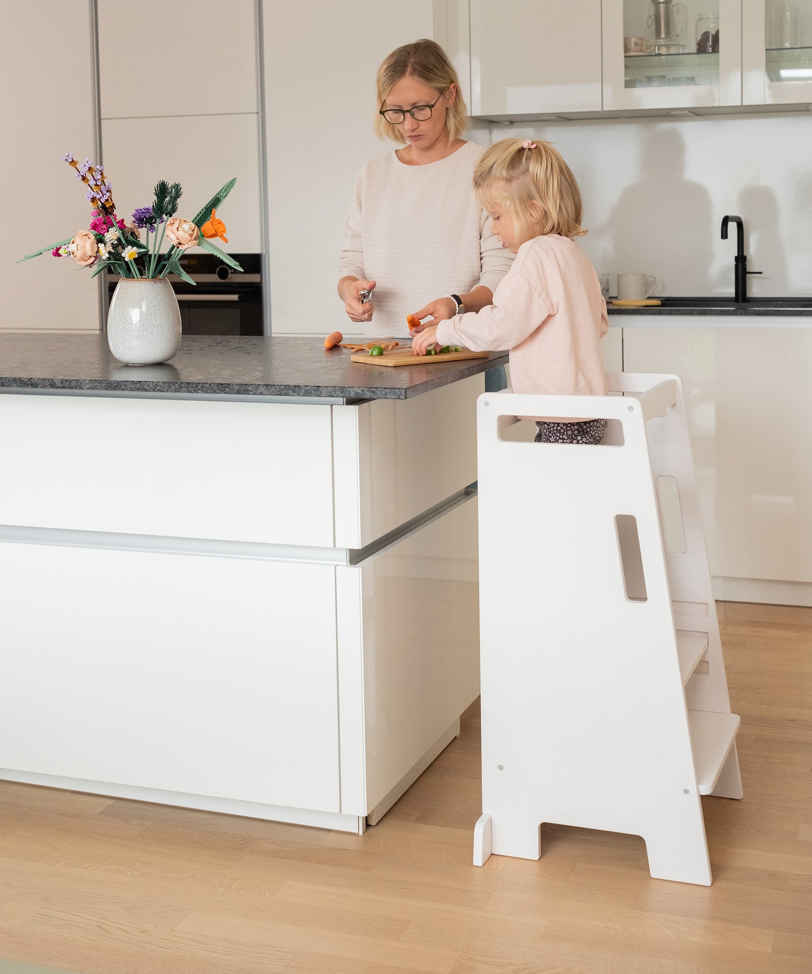 Gemeinsam lernen*Mit dem Lernturm nimmt dein Kind aktiv am Familienalltag teil.
Darin steht es Seite an Seite mit dir und kann spielerisch lernen.|Ein Kind steht auf einem Lernturm und hilft einer Erwachsenen beim Schneiden von Gemüse.