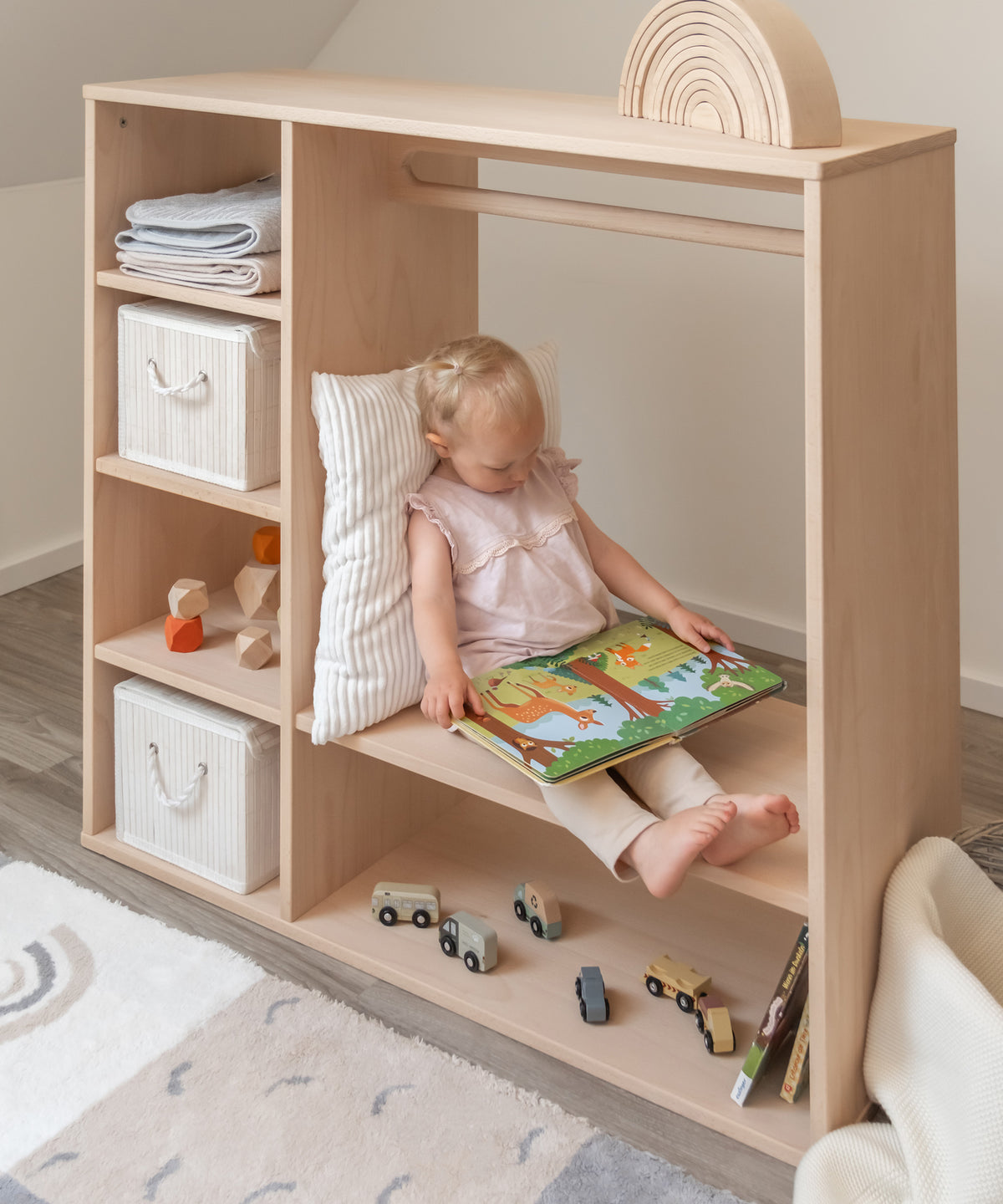 Kleines Mädchen sitzt auf großem Regalboden im Montessori Kleiderschrank und schaut ein Buch an.