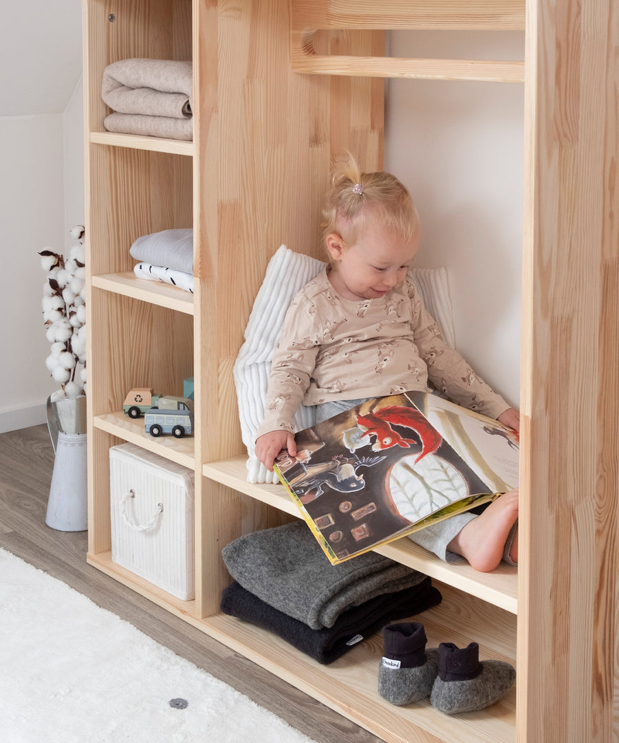 Kindgerecht & vielseitig*Der Montessori Kleiderschrank kann auch als gemütlicher Ort zum Lesen oder Spielen genutzt werden.|Kleines Mädchen sitzt auf großem Regalboden im Montessori Kleiderschrank und schaut ein Buch an.