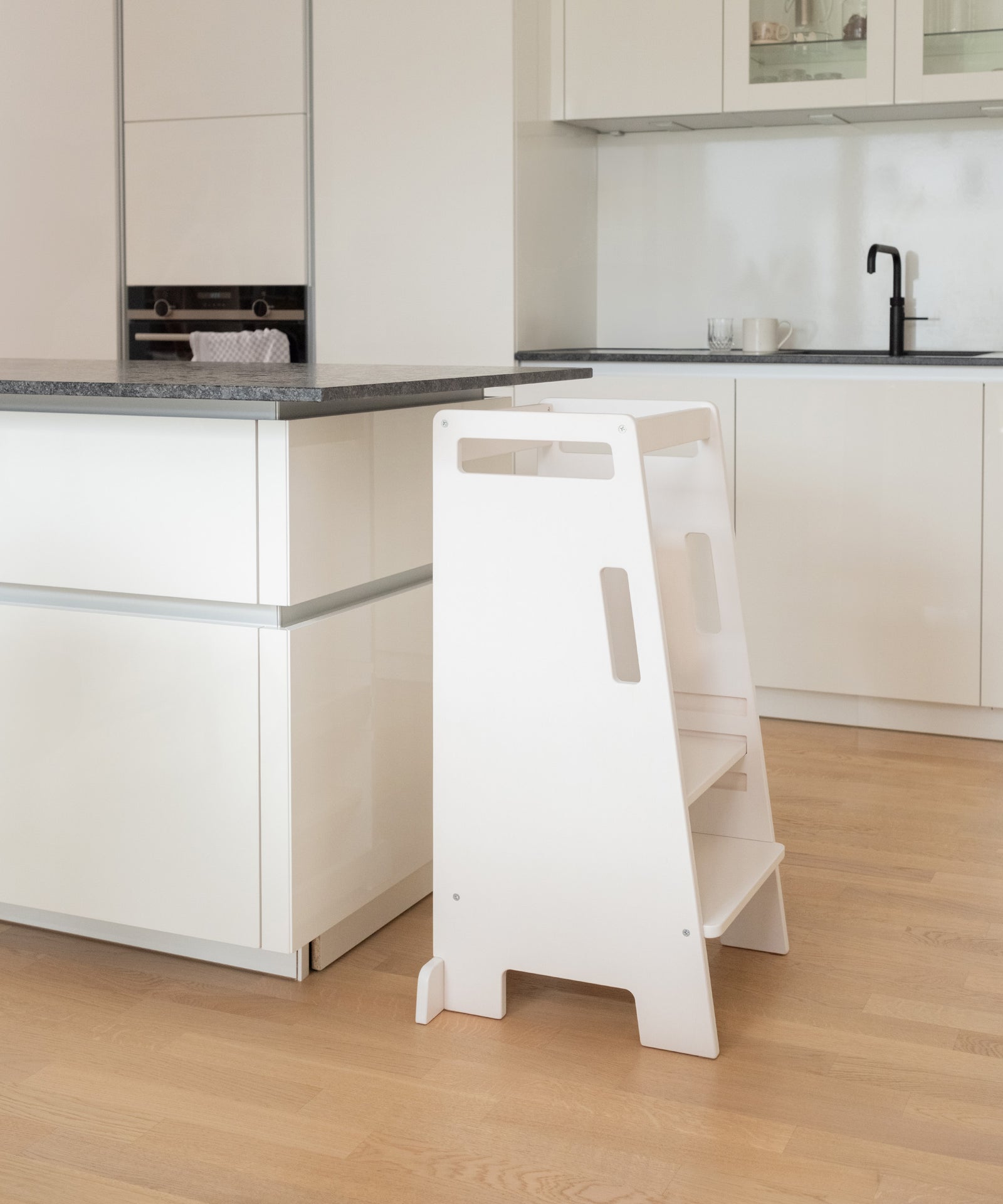 Von Montessori inspiriert*Mit dem Montessori-Möbelstück kann dein Kind Küche, Esstisch oder Waschbecken sicher erreichen und Neues entdecken.|Ein weißer Lernturm steht in einer Küche vor einer Kücheninsel.