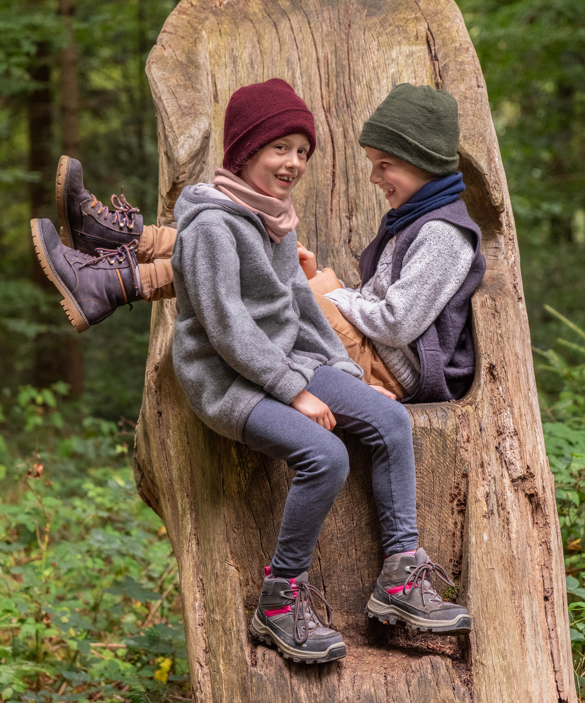 Zwei Kinder mit Schlauchschal und Strickmütze sitzen auf Baumstumpf im Wald.