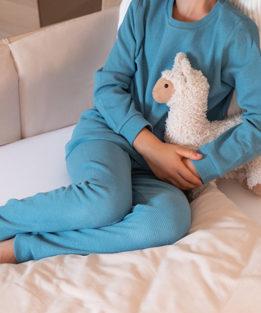 Waffelstruktur*Die leicht strukturierte Oberfläche ist besonders saugfähig, herrlich weich und angenehm zu tragen.|Kind mit Schlafanzug Blau Waffel sitzt mit Kuscheltier auf Bett.