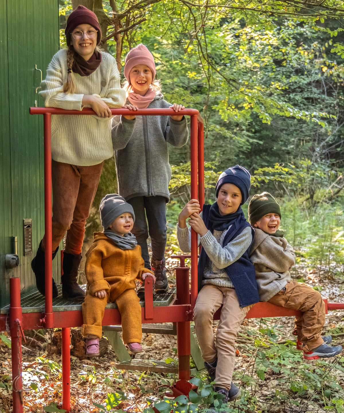 Fünf Kinder unterschiedlichen Alters mit Strickmützen in fünf verschiedenen Farben auf Bauwagen im Wald.