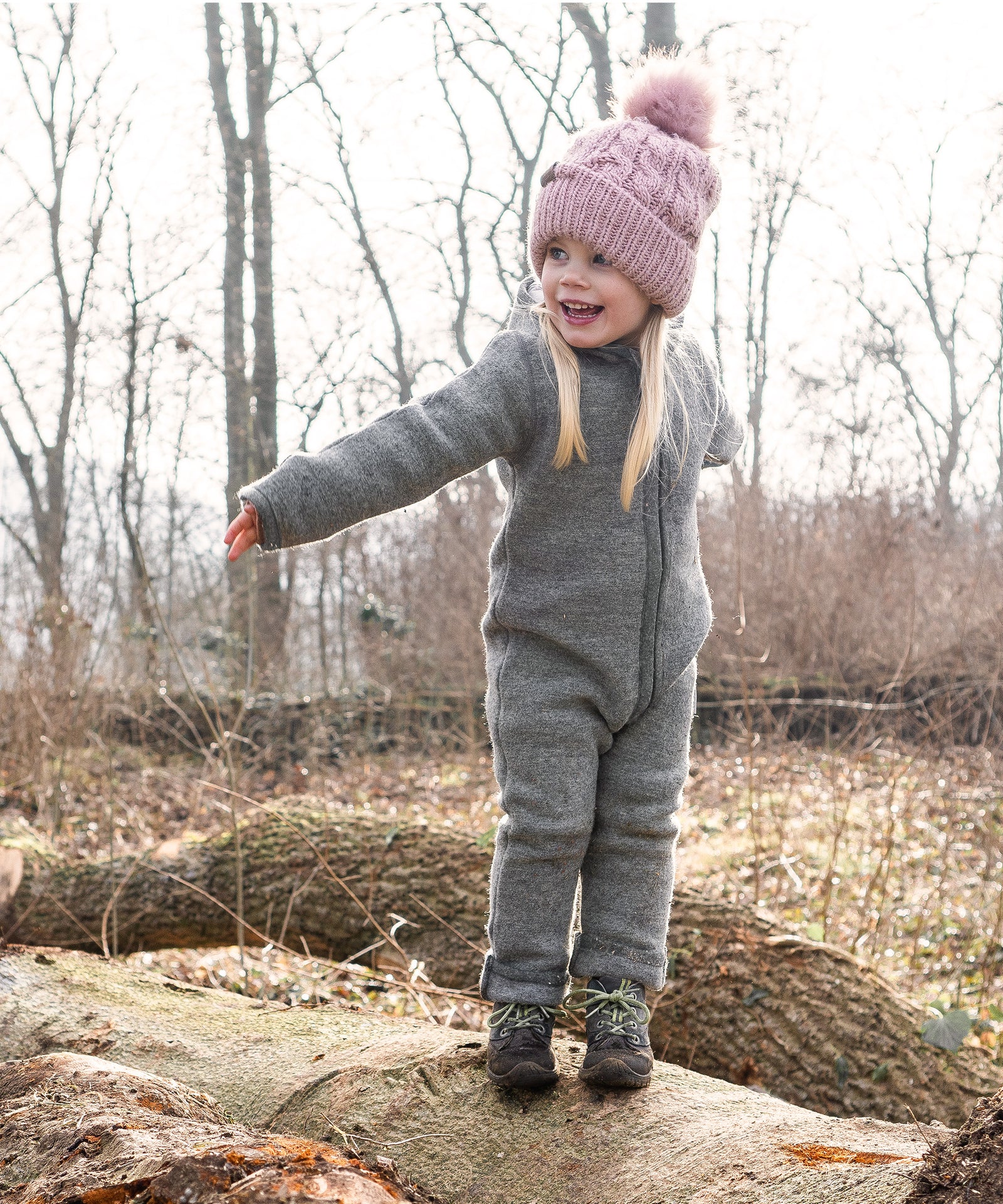 Reine Schurwolle*Die eingesetzten Walkstoffe sind zu 100 % ein reines Naturprodukt, es werden keinerlei chemische Fasern verwendet.|Mädchen mit Walkoverall steht auf Baumstamm im Wald.
