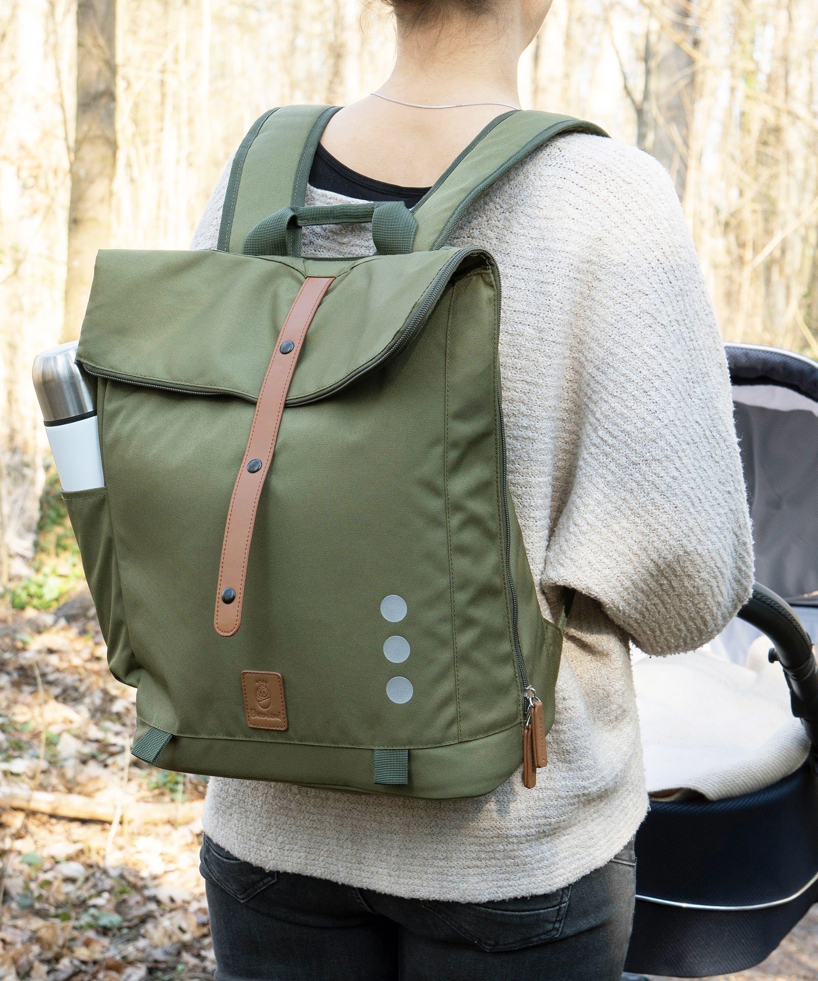 Recyceltes Polyester*Der Wickelrucksack in zwei Unisex-Farben wird nachhaltig unter fairen Bedingungen aus recycelten PET-Flaschen hergestellt.|Frau mit Wickelrucksack Olivgrün auf dem Rücken geht mit Kinderwagen spazieren.