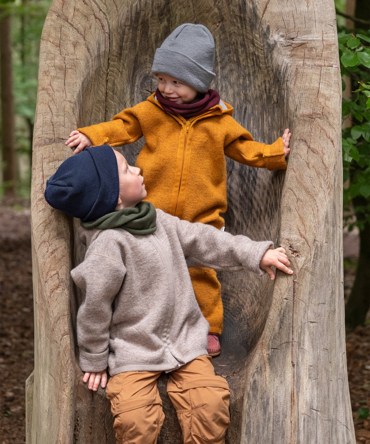 Zwei Kinder mit Strickmützen auf Baum im Wald.