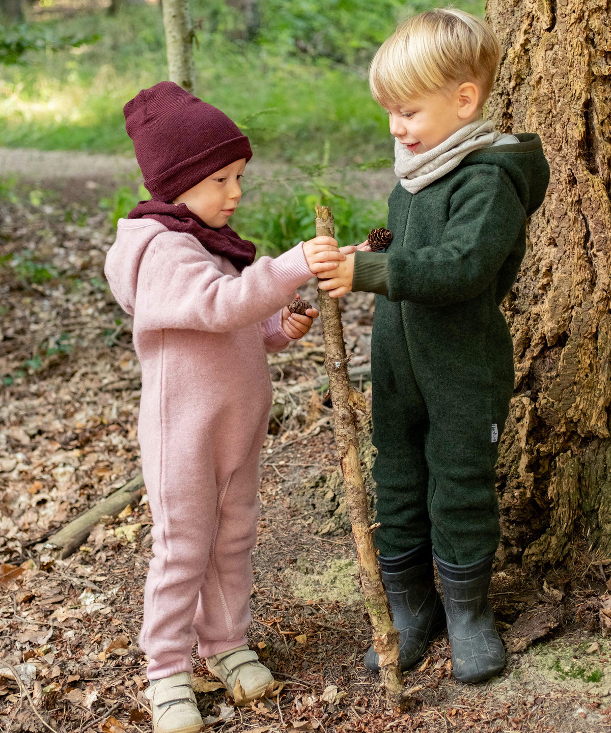 Zwei Kinder mit Wollfleeceoveralls spielen im Wald.