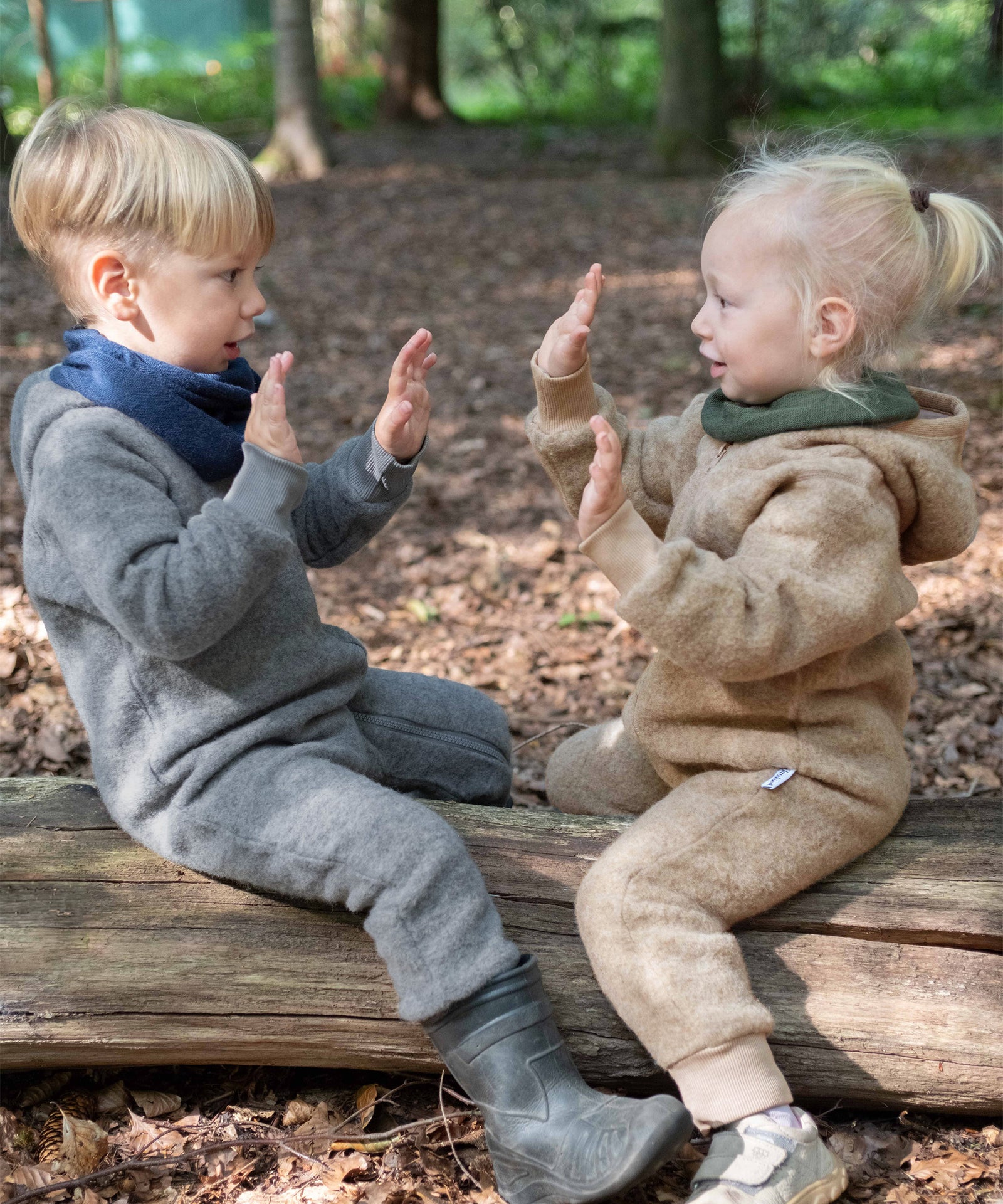 Zwei Kinder mitt Wollfleeceoverall auf Baumstamm im Wald.