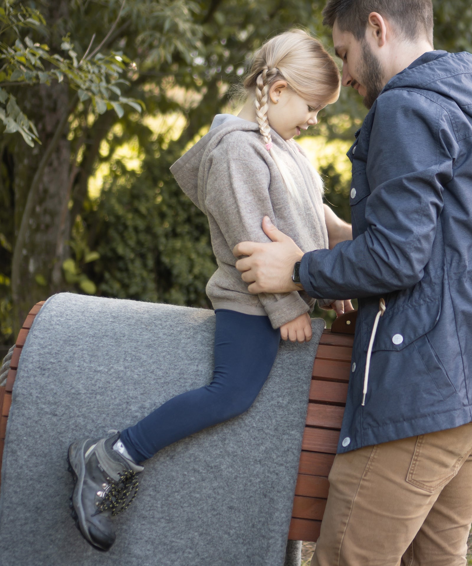 Vielseitig & robust*Ob drinnen oder draußen - an kühlen Tagen schenkt unsere Walkdecke deinem Kind Wärme, Schutz und Geborgenheit.|Mädchen sitzt auf einem Holzpferd. Unter ihr liegt zum Schutz eine Walkdecke. Ihr Papa steht daneben und hilft ihr.
