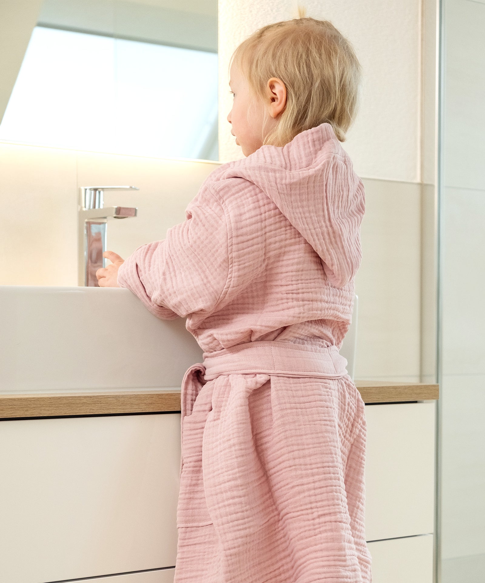 Kind mit Bademantel Musselin mit Kapuze steht am Waschbecken.