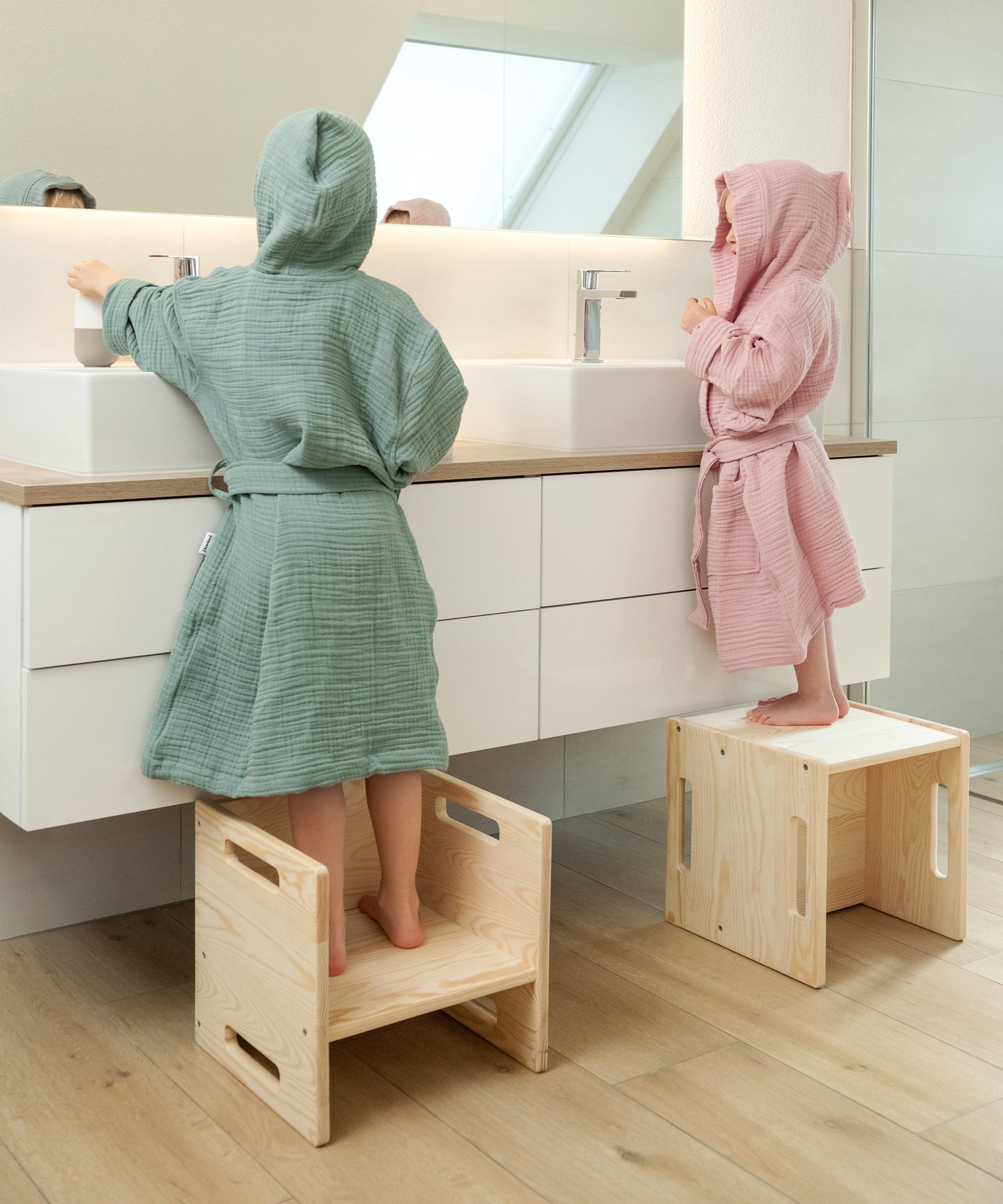 Perfekte Passform für jedes Alter*Ob fixierter Gürtel bei kleinen Größen oder lose mit Schlaufen ab Größe 110/116 - der Schnitt wächst einfach mit.|Zwei Kinder mit Bademantel aus Musselin auf Hockern vor dem Waschbecken.