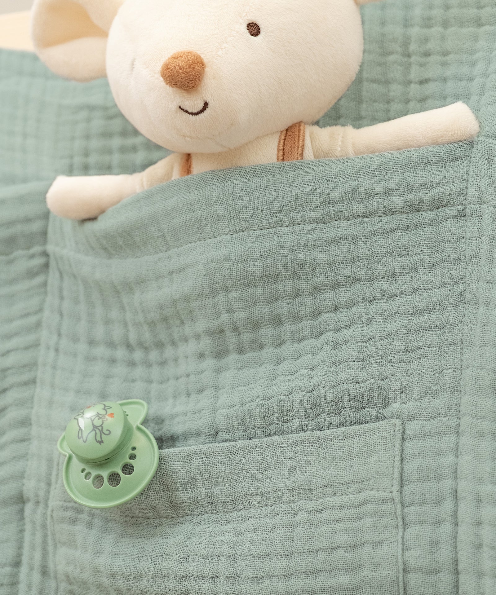 Hochwertige Materialien*100% Bio-Musselin macht den Organizer hautfreundlich, langlebig und dank OEKO-TEX® Zertifizierung ideal für Kinder.|Nahaufnahme eines grünen Bett-Organizers mit Kuscheltier und Schnuller in den Taschen.