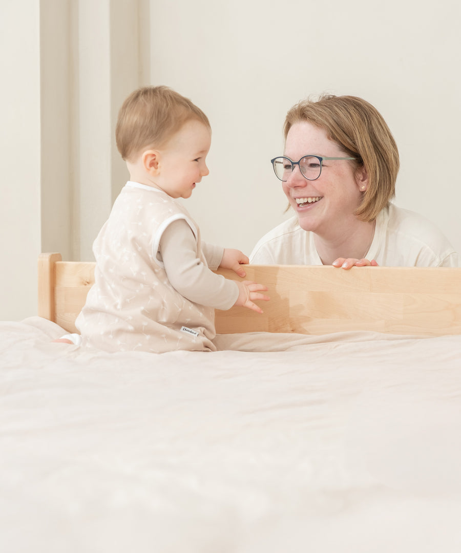 Baby sitzt auf dem Bett und hält sich an dem Rausfallschutz fest.