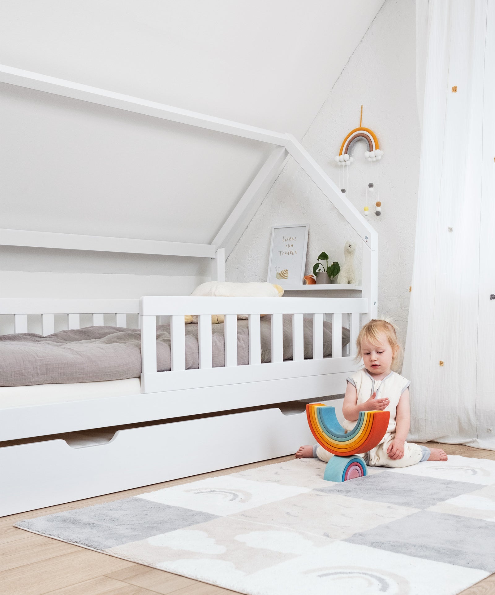 Abnehmbarer Rausfallschutz*Mit dem abnehmbaren Rausfallschutz schläft dein Kind von Anfang an sicher und geschützt.|Mädchen sitzt vor Hausbett Kiefer weiß mit Bettkasten auf dem Teppich und spielt.