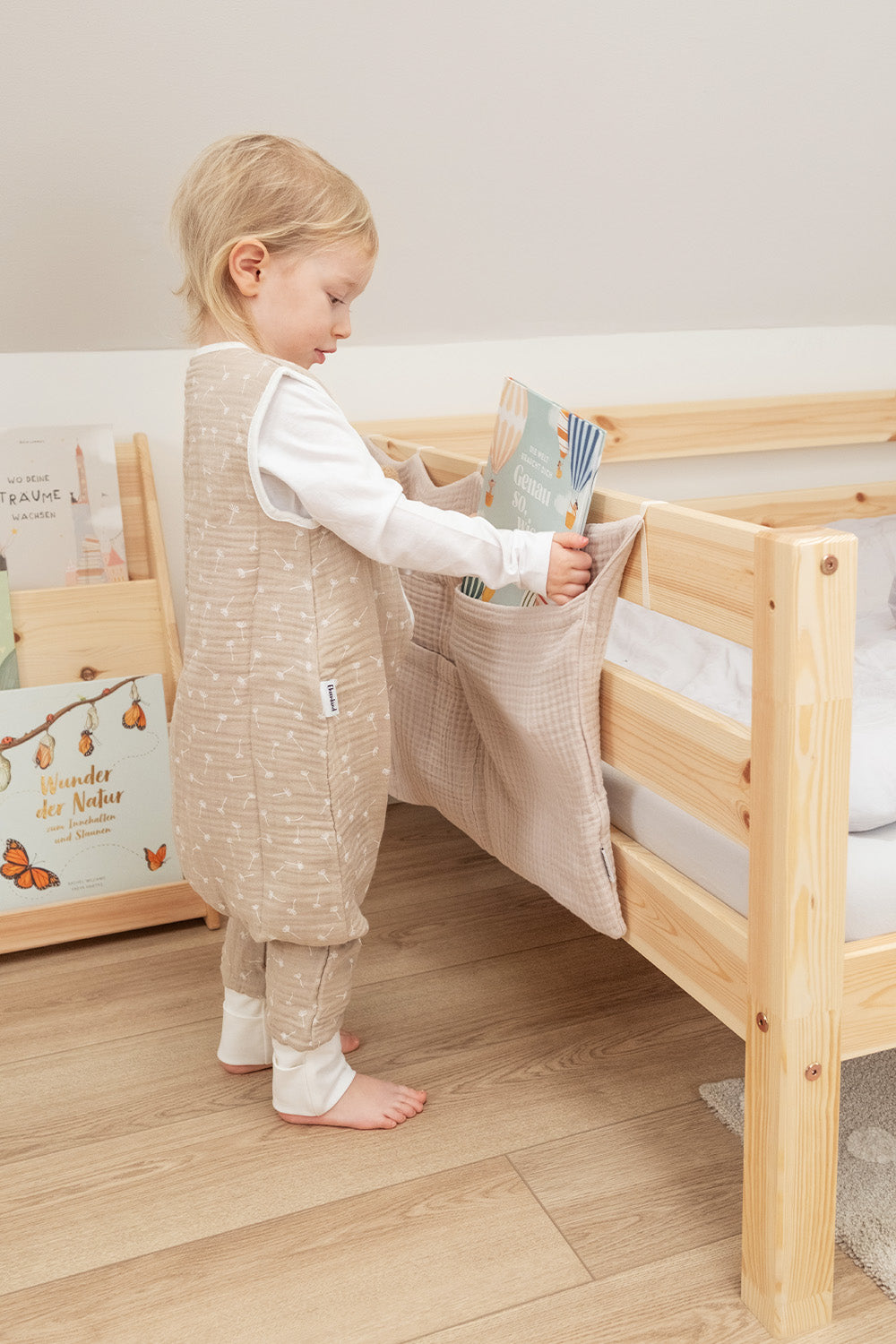 Kind in Musselin-Sommerschlafsack mit Beinen steht vor Bett.