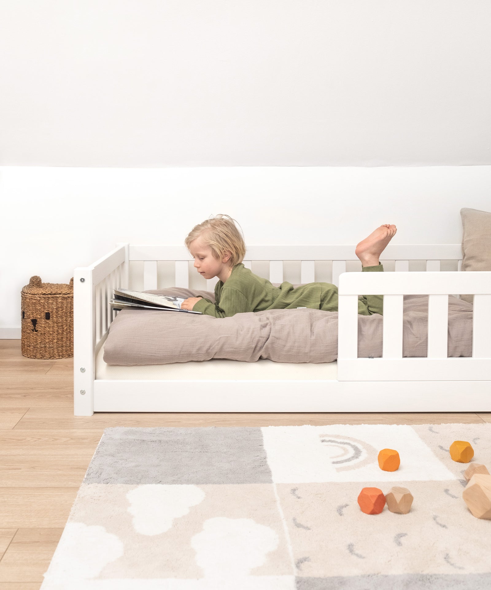 Kind liegt in Kinderbett Bodentief aus weißem Kiefernholz und schaut Buch an.