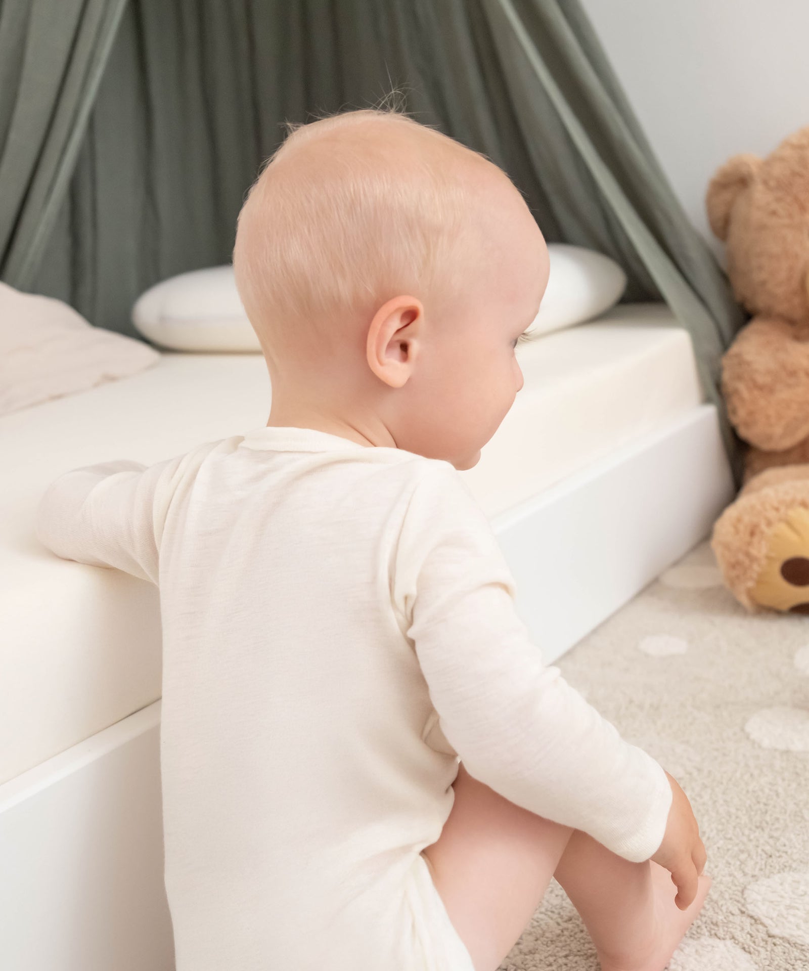 Selbstreinigende Wirkung*Wolle nimmt kaum Gerüche auf - Bakterien haften schlecht an, Gerüche werden reduziert. Daher ist kein häufiges Waschen nötig.|Baby sitzt neben Bett.