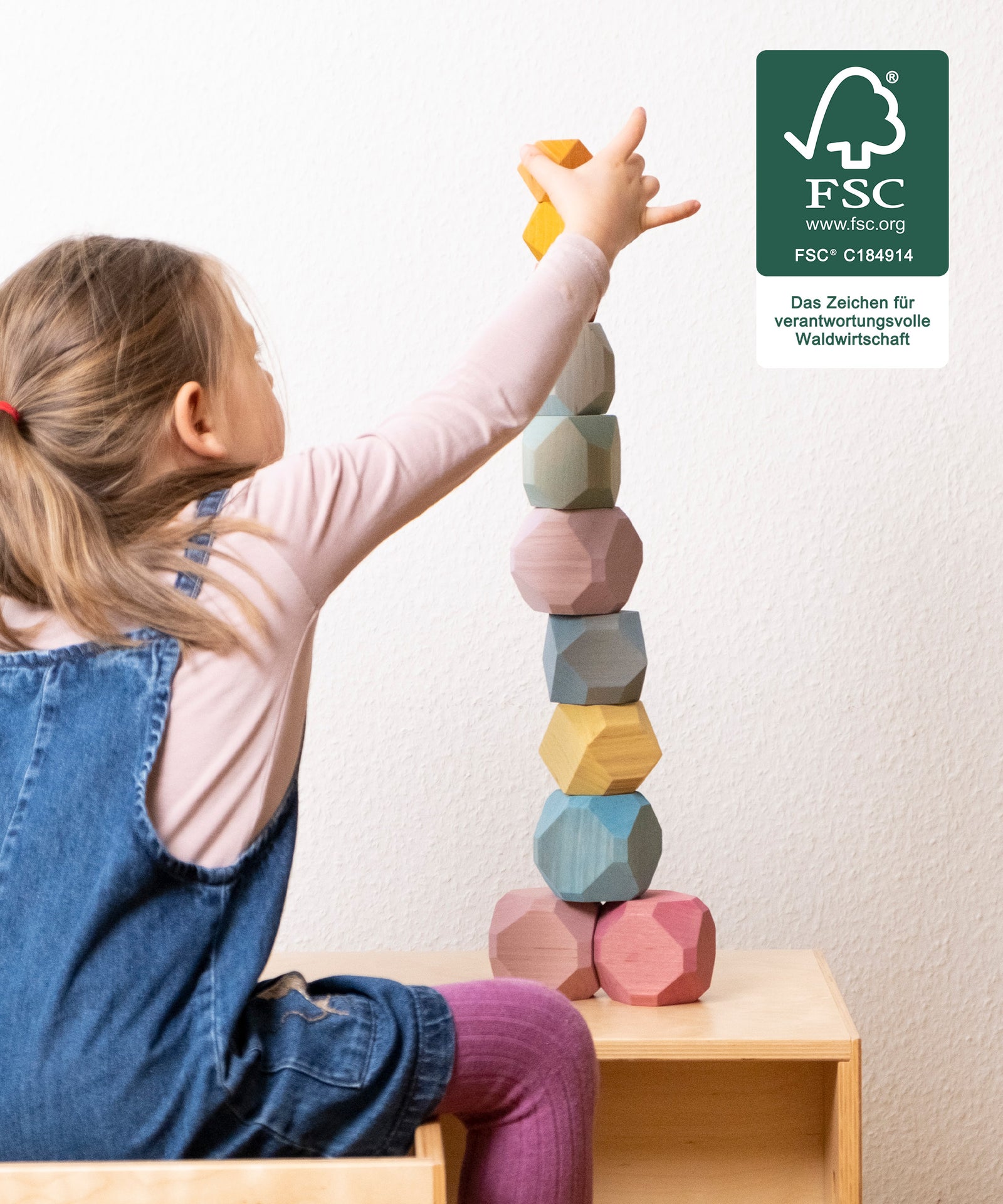 Fördert Motorik & Vorstellungskraft*Bauen und Stapeln schulen spielerisch Feinmotorik, Hand-Augen-Koordination und das Vorstellungsvermögen.|Mädchen baut Turm aus Balanciersteinen zum Stapeln. FSC Logo auf Foto.