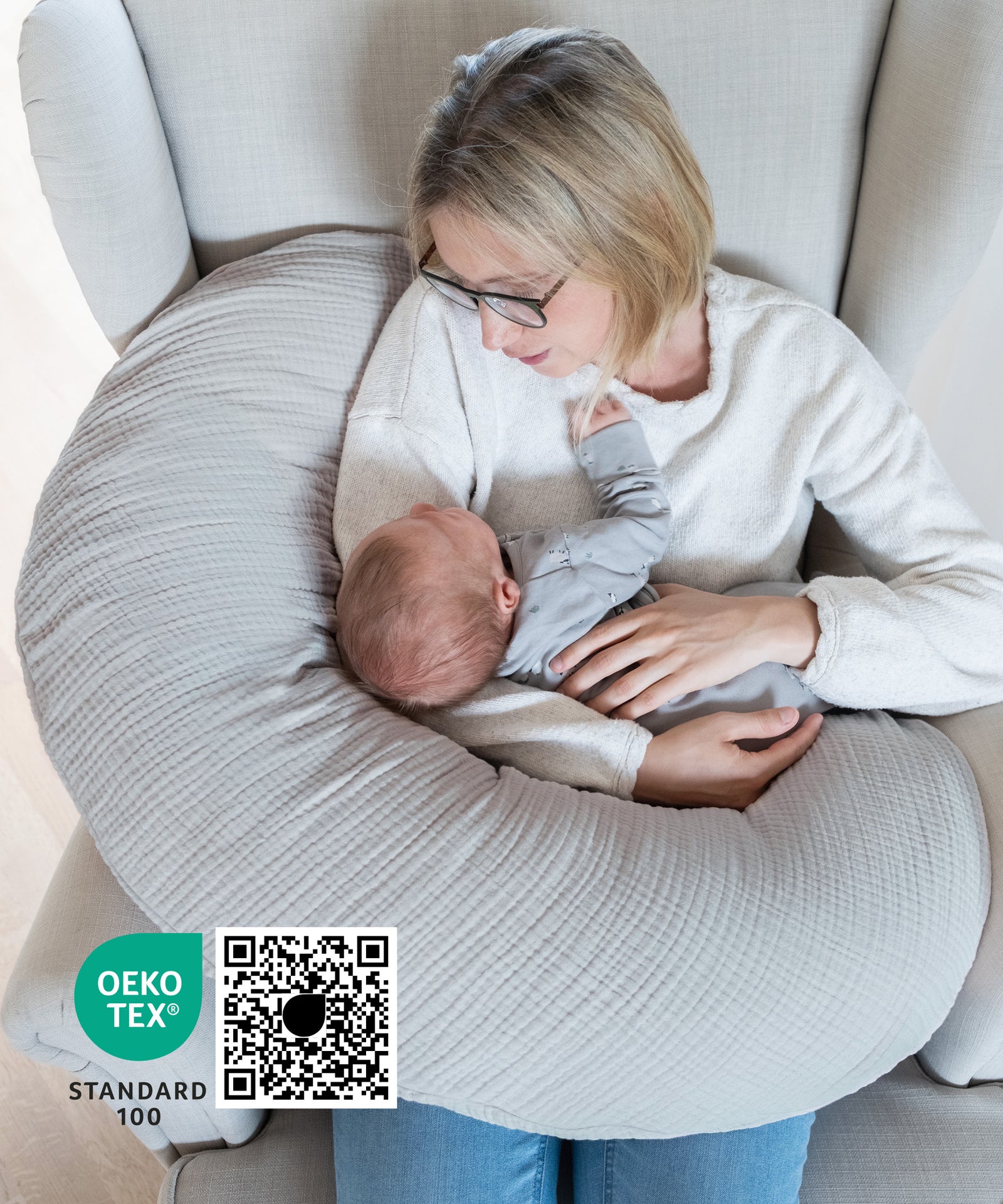 Optimale Unterstützung beim Stillen*Indem das Stützkissen das Gewicht des Babys abfedert, wird die Belastung von Armen, Schultern und Nacken reduziert.|Frau mit Baby sitzt mit Stillkissen Musselin in Sessel, Stillkissen Oekotex zertifiziert..