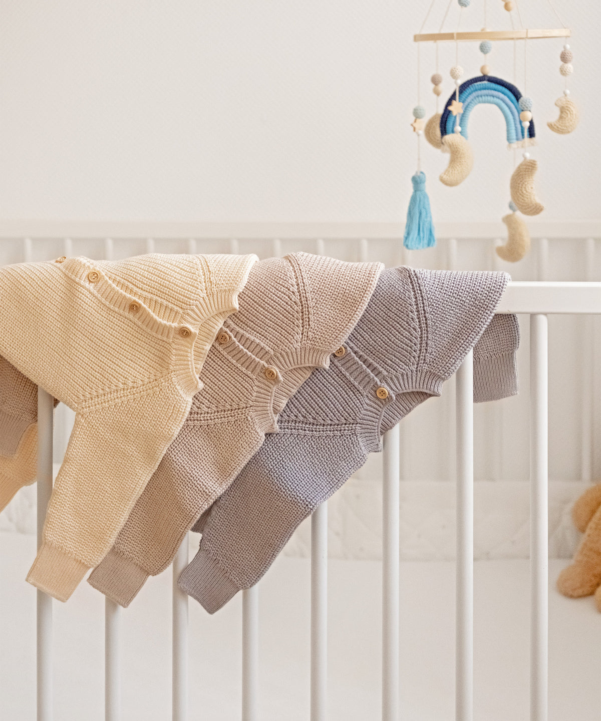 Strickoveralls in Beige, Natur und Hellgrau aus Bio-Baumwolle, dekorativ über der Babybett-Stange aufgehängt.