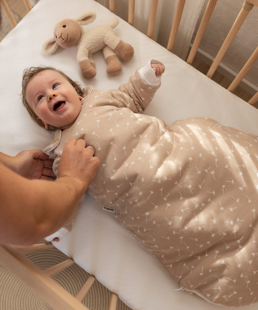 Wohlige Wärme für kalte Nächte*Mit 3.5 TOG sorgt der Babyschlafsack für Wärme, Geborgenheit und ruhigen Schlaf – ideal für Raumtemperaturen von 13-16 °C.| Baby trägt den 3.5 TOG Schlafsack in Beige Pusteblume und liegt im Babybett.
